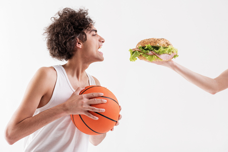 Starving skinny male basketball player is opening his mouth widely while looking at appetite sandwich in female arm. Isolatedの写真素材