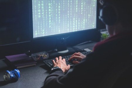 Decoding information. Back view of involved experienced hacker is sitting in dark lighted room and working on computer while typing on keyboardの写真素材