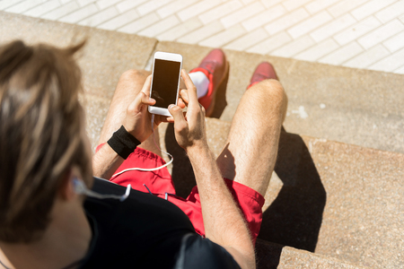 Top view of young man in sportswear sitting on stairs outside. He is holding mobile and listening to music via earphones. Focus on smartphoneの写真素材