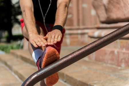 Close up of hands and leg of youthful athlete doing stretching exercises on stairs with railing. He is listening music via earphones. Focus on metal handholdの写真素材