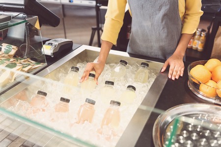Cool down. Close-up top view of arms of friendly female worker. She is standing near refrigerator and taking bottle of juice from box with ice in organic storeの写真素材