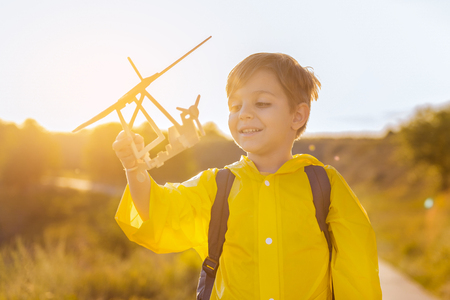 Portrait of happy child holding wooden aircraft and laughing. He is standing with backpack on road near grass fieldの写真素材