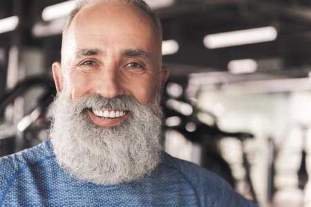 Feeling happy. Close-up portrait of cheerful old man with beard. He is looking at camera with smile while standing in gym. Copy space in the right sideの写真素材