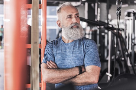 Strong and confident. Stylish old man with beard is leaning on shelf in modern gym and looking aside thoughtfully while standing with crossed arms. Portraitの写真素材