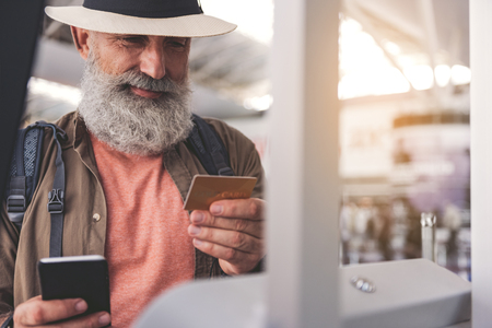 Portrait of outgoing bearded old man watching at credit card while keeping mobile in hand near terminalの写真素材