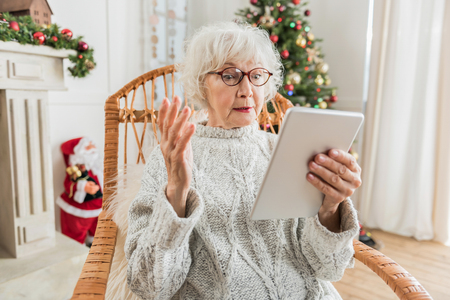 How to use. Senior woman is holding tablet and looking at screen while expressing discontent and perplexity. Christmas tree and Santa Claus are on backgroundの写真素材