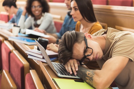 Bored young male student is sleeping on lecture. He is sitting at table and leaning head on laptop keyboardの写真素材