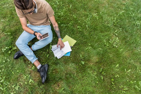 Top view of concentrated male student doing homework in the nature. He is sitting on lawn and...