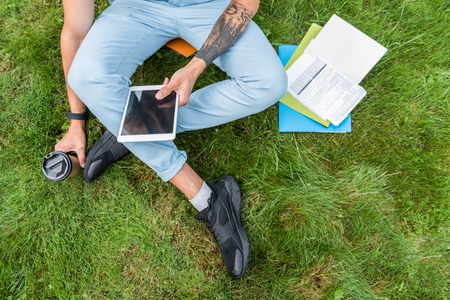 Top view close up of male legs sitting on meadow grass near learning material. Man is holding cup of coffee and gadget. Tattoo on his armの写真素材