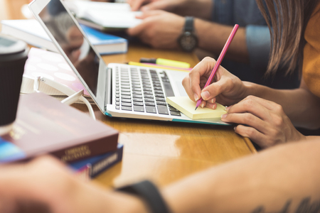 Close up of female hands making notes on sticker. Girl is sitting at desk near laptop next to her group matesの写真素材