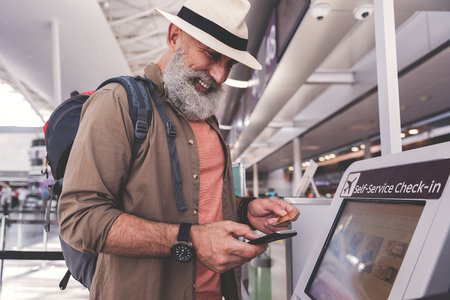 Cheerful bearded old man writing in mobile while keeping credit card in hand. He is standing in airportの写真素材