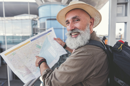Cheerful mature bearded tourist is pointing on certain place at unfolded map. He looking ahead with...