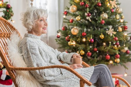 Feeling comfortable. Elegant old woman in cozy chair is enjoying hot tea while sitting with Christmas tree on background. Copy space in the right sideの写真素材