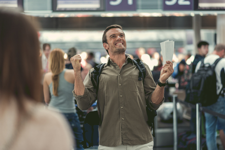Extremely happy. Selective focus of delighted adult man is standing at international airport and expressing gladness while holding two tickets on plane. People on backgroundの写真素材