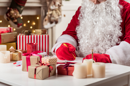 Close up of concentrated old man with big white beard writing list of children in notebook. He is sitting in Santa costume. Focus on gift boxes on tableの写真素材