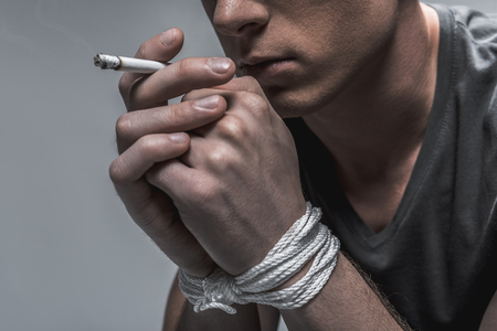Realizing problems with self-control. Close up of pensive young man smoking cigarette. His hands are tied up with a rope. Isolated and copy space in left sideの写真素材