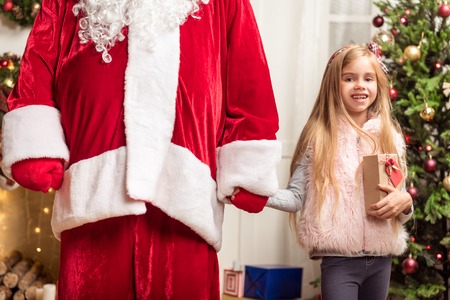 Portrait of happy girl standing and holding hand of Santa Claus. She is holding gift box and looking at camera with excitementの写真素材