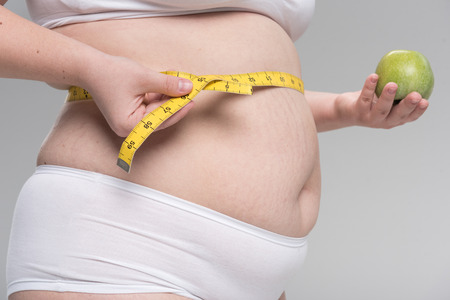 I must go on a diet. Close up of large abdomen of young fat woman measuring her waist by tape. She is standing and holding an apple. Isolatedの写真素材