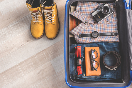 Camera and other accessories are locating on clothes in open suitcase. Boots near luggage at floor. Top view close up. Copy space on left sideの写真素材