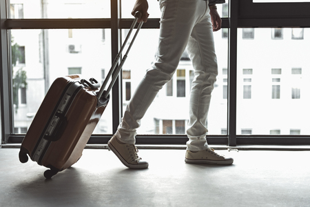 Ready for trip. Close-up of legs of stylish cute man who is carrying suitcase while going along big window with cityscapeの写真素材
