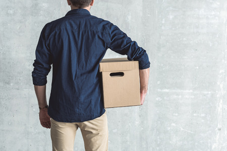 Delivery concept. Back view of stylish courier is holding cardboard box. He is standing against gray wall background. Copy space in the right sideの写真素材