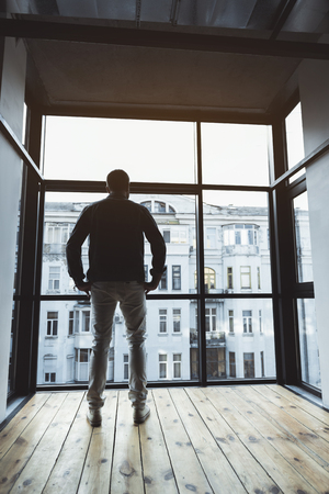 Great view. Low angle full length of thoughtful trendy man who is standing against big window and looking through glass on cityscape. Copy space in the right side. Back viewの写真素材
