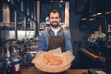 Portrait of cheerful unshaven worker holding croissant on salver in confectionary shop. Occupation...