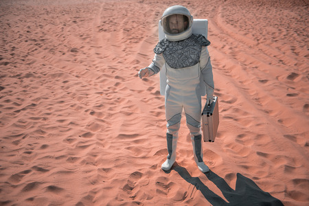 Serious spaceman wearing white armor is standing on red sand under hot sun and rising hand. He...