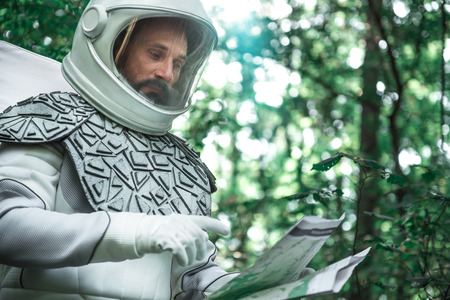 Concentrated cosmonaut wearing white helmet is reading unfolded papers while standing in forest. Low...