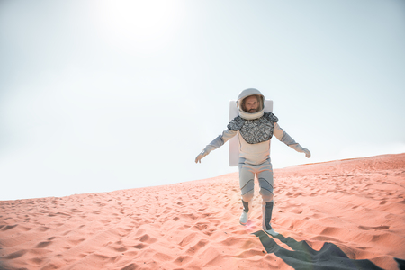 Serious spaceman wearing white armor is hardly trying to move forward on desert and looking at camera with determination. Full length portrait. Copy space on left sideの写真素材