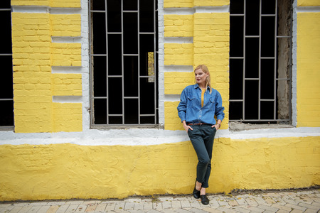 Full length portrait of confident blond girl standing near yellow building outdoor and posing. Copy spaceの写真素材