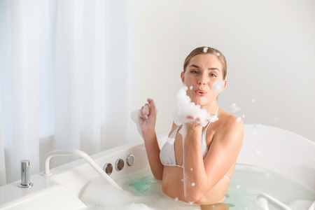 Portrait of playful young woman blowing foam forward as if she is sending air kiss. She is sitting...