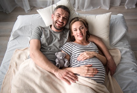 Top view of excited future parents lying on bedding and hugging. They are looking at camera and smiling. Expectant lady is touching her belly with fondnessの写真素材