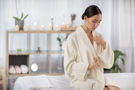 Pleasant weekends. Attractive relaxed asian girl is sitting on massage table at wellness center with candles in background. She is taking off her white bathrobe. Copy space in the left sideの写真素材