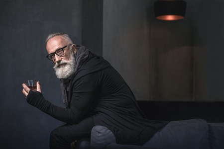 Portrait of smiling bearded pensioner tasting delicious beverage while situating in room. Relax concept. Copy spaceの写真素材