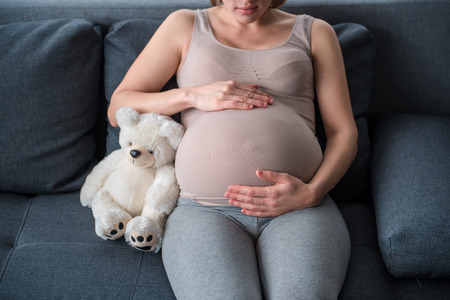 Focus on belly of pregnant woman. She is sitting on couch near teddy bear and embracing tummy by...