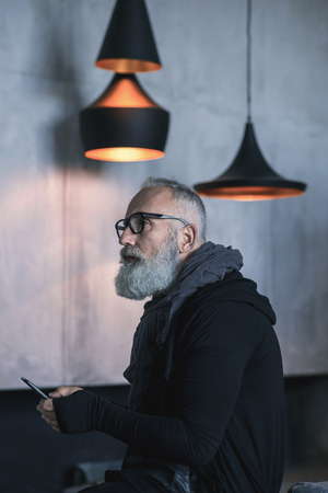 Side view serene bearded old man typing in mobile while situating on soft sofa. Technology conceptの写真素材