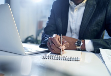 Analyzing important information. Close-up of hands with pen of young professional african manager who is sitting at table with modern laptop while taking notes in his copybook. Selective focusの写真素材