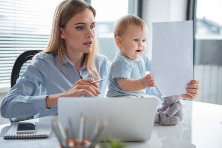 Portrait of uneasy mother and happy kid looking at paper while holding it in hand in modern office. Job and baby conceptの写真素材
