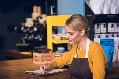Side view smiling worker writing information while locating at counter in modern cafe. Labor concept