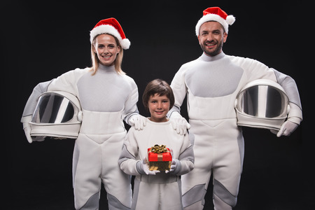 Christmas in space concept. Portrait of cheerful family of astronauts in red hats are standing and holding helmets while looking at camera with smile. Cute boy is standing with gift box. Isolatedの写真素材