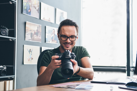 Portrait of cheerful young unshaven man looking at gadget for taking photos while situating at table in apartment. Occupation conceptの写真素材