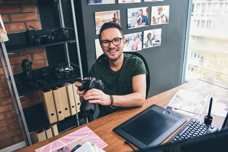 Portrait of beaming unshaven man keeping appliance for taking photos while sitting at table in room. Occupation conceptの写真素材