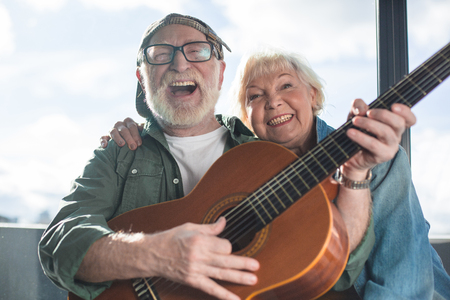 True happiness. Portrait of two sincere pensioners sitting together. Woman is cuddling man in cap while he is playingの写真素材