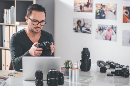 Portrait of serious bearded young retoucher looking at camera while holding it. He working at table. Job conceptの写真素材