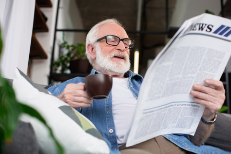 Funny story. Low angle portrait of bearded smiling pensioner reading journal relaxing on sofa with hot coffeeの写真素材
