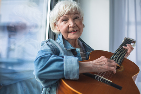 Waist up portrait of senior lady holding musical instrument and looking at camera. She is smiling while sitting at windowの写真素材