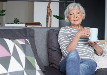 Portrait of old mature lady sitting on couch and smiling. She is enjoying tasty drink while relaxing at living room. Copy space in left sideの写真素材