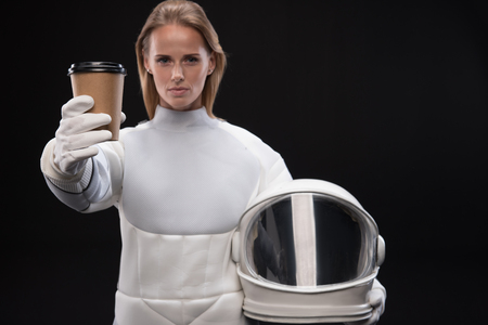 Serious young trained female cosmonaut is stretching coffee to camera while holding helmet and standing confidently. Isolated background. Selective focusの写真素材
