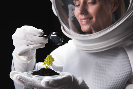 Cheerful female cosmonaut researcher wearing helmet is standing and looking through magnifier on green plant on her palm while expressing gladness and curiosity. Selective focus. Isolated. Close-upの写真素材